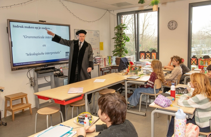 Bart Schermer wearing a professor’s gown and pointing to an electronic whiteboard. Children sit at their desks and listen.