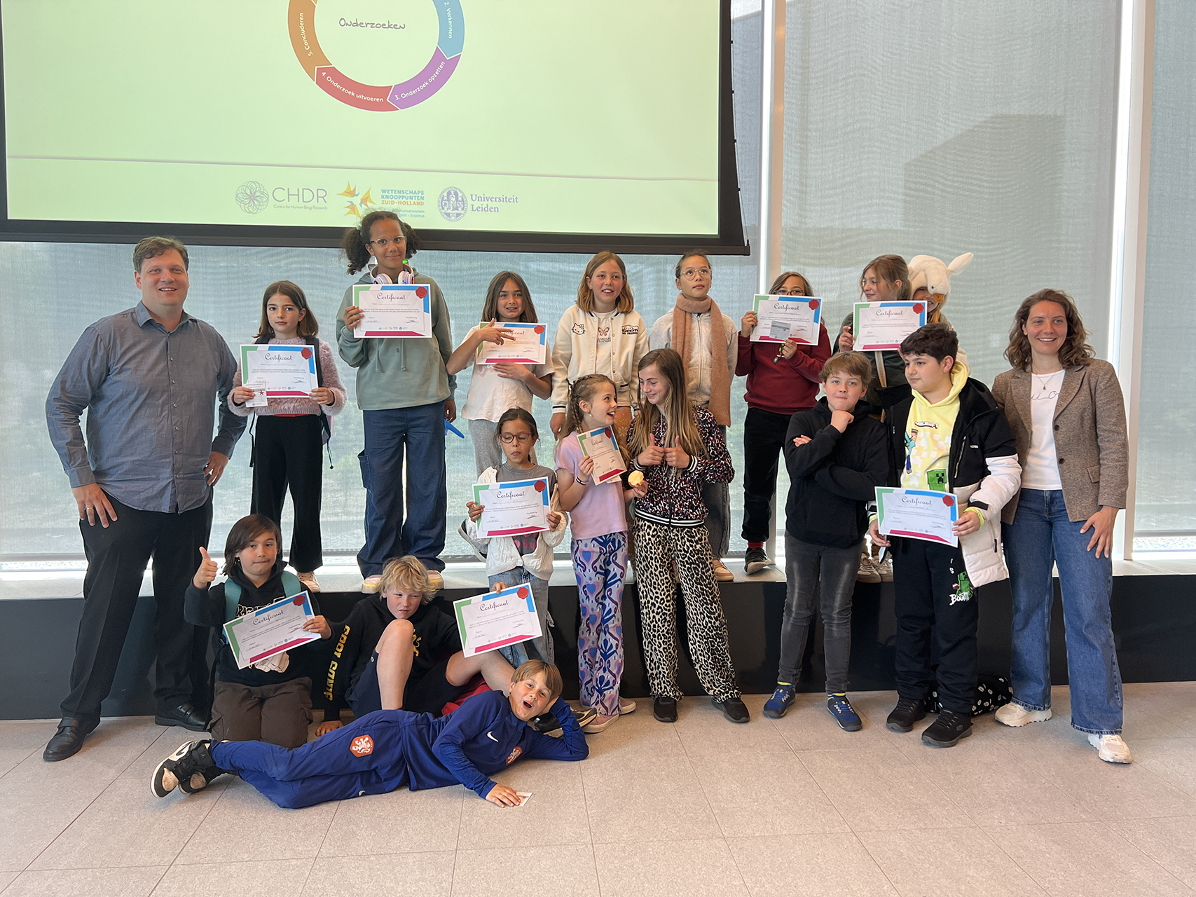A man and a woman pose with a group of children happily displaying their certificates.