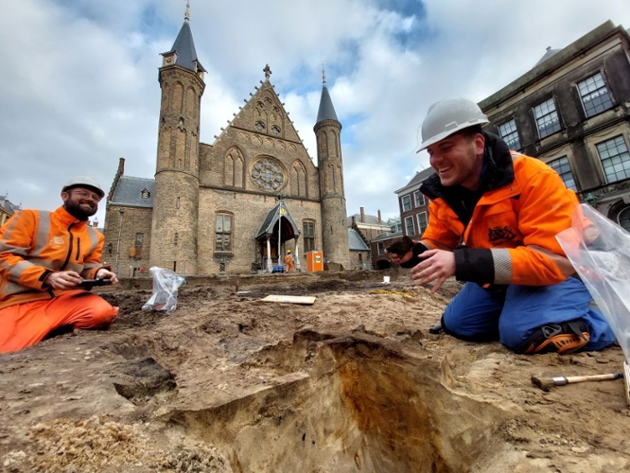 Two men in orange hi-vis workwear and helmets kneel in the sand in front of Ridderzaal at Binnenhof, The Hague.