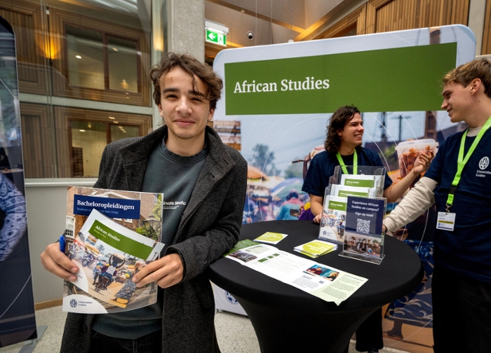 A lad with brown hair wearing a long grey coat stands in front of an African Studies banner.