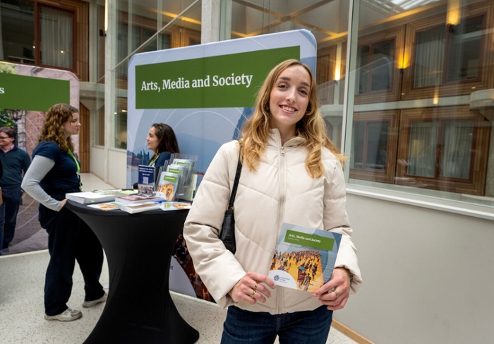 A girl with long blonde hair and wearing a white jacket holds up an Arts, Media and Society brochure.