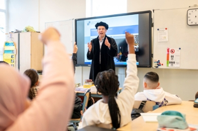 A professor in gown stands in front of a class of children with their hands raised.