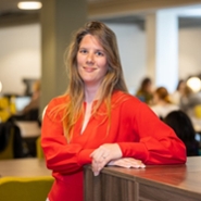 Een vrouw in een oranje bloes leunt op een tafel en kijkt in de lens