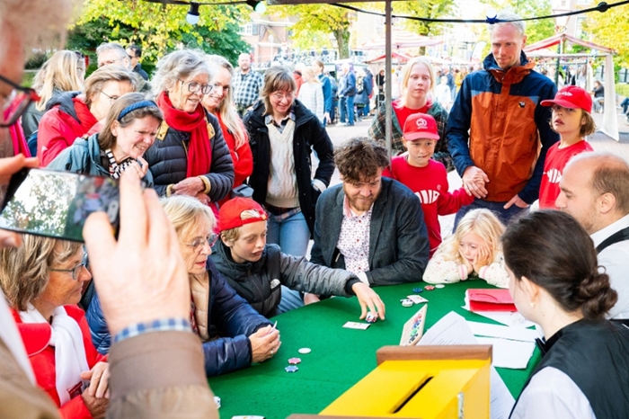 Een groep mensen staat bij een tafel waaraan andere mensen een kaartspel spelen