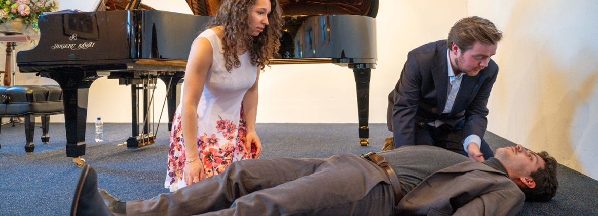 Mannelijke student ligt op het podium, mannelijke en vrouwelijke student buigen er bezorgd overheen met piano op de achtergrond.