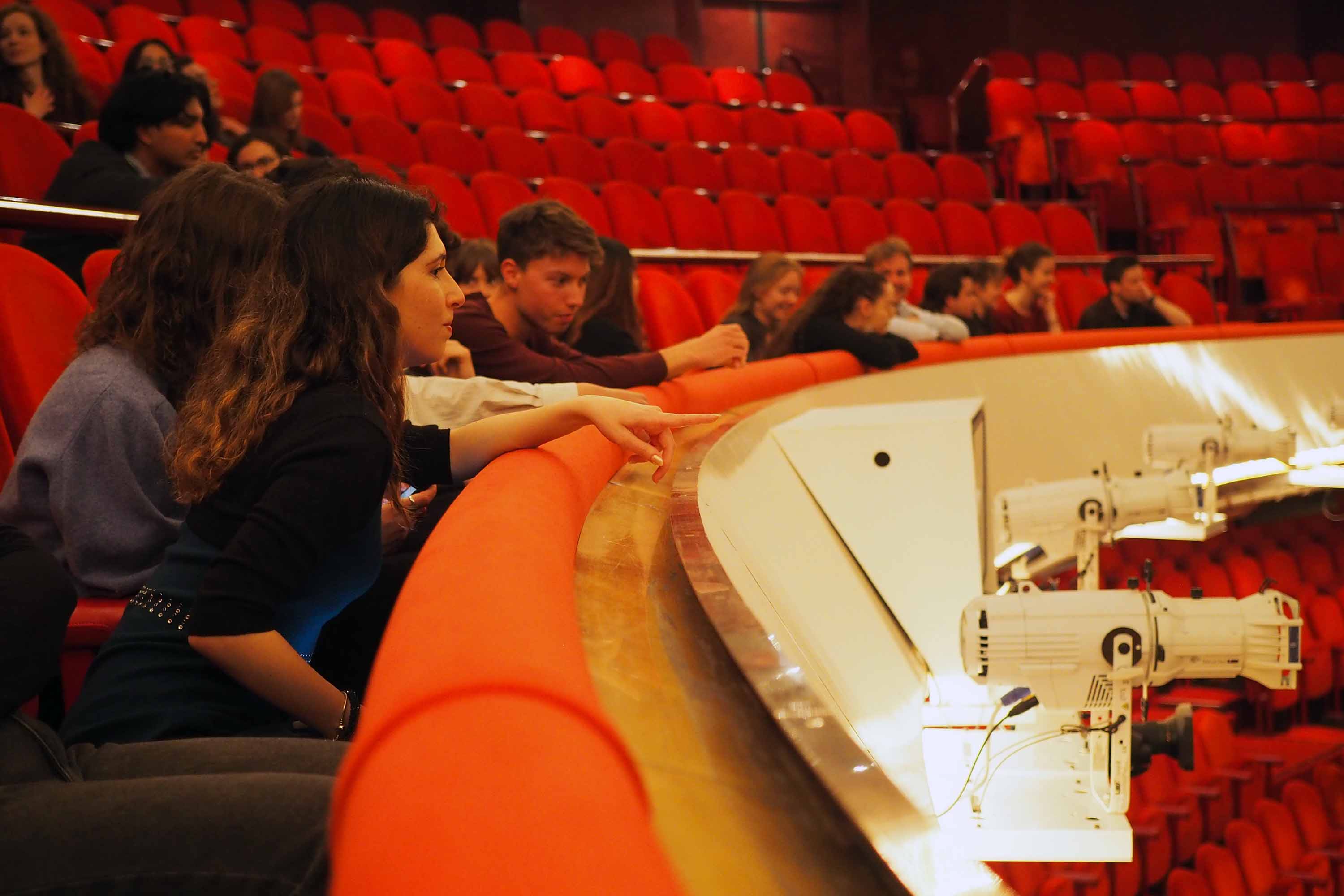 In een operazaal vol rode stoeltjes wijst een student richting het podium.