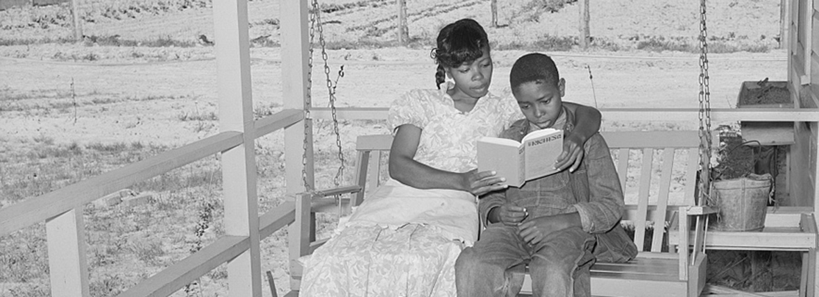 Echtgenote van FSA (Farm Security Administration) leest een boek voor aan haar zoon op een schommel op de veranda. Een boeren tuin op de achtergrond. Sabine Farms, Marshall, Texas.