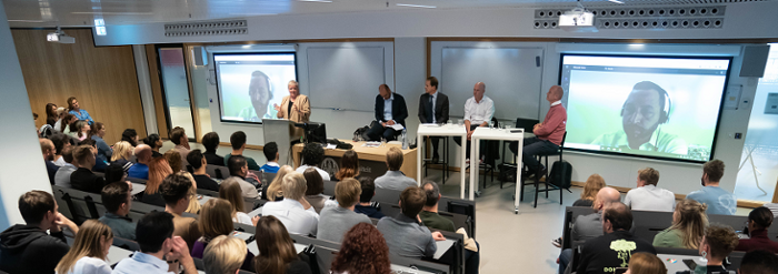 Foto van de zaal in Wijnhaven waar het debat plaatsvond. Het publiek zit in de zaal, voorin de panelisten waarvan een via videobeeld op een scherm.