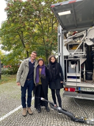 In the photo, three people are standing next to an open service vehicle equipped with technical equipment. Inside the vehicle, cable reels and mechanical components are visible, intended for sewer inspection or maintenance. On the ground lies a large cylindrical device that appears to be an inspection robot, connected to the vehicle by a cable.