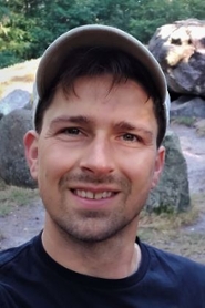 Een glimlachende man met stoppelbaard en honkbal pet. Op de achtergrond een reusachtige kei.